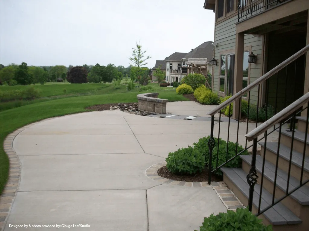 A concrete patio with a stone wall feature, leading to a lawn and trees, viewed from beside a house staircase.