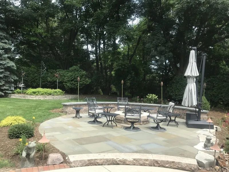 A stone patio with a central fire pit surrounded by outdoor chairs, a closed umbrella, and a stone retaining wall.