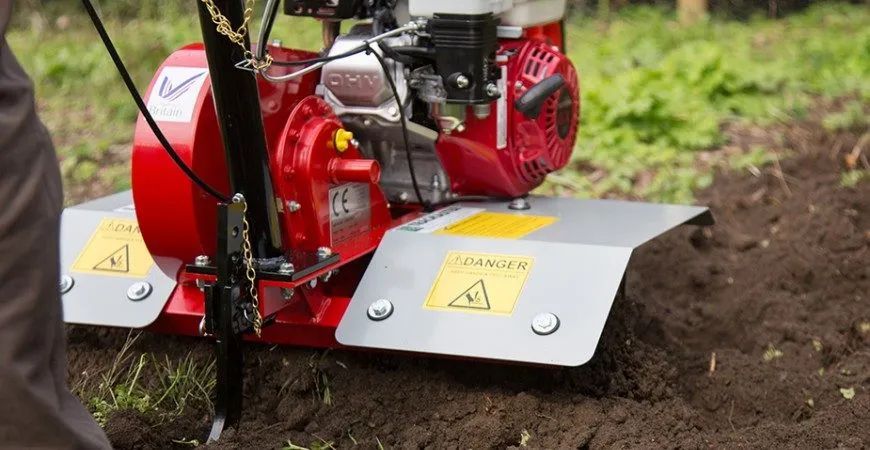 A person uses a red motorized garden tiller to turn soil in a field.
