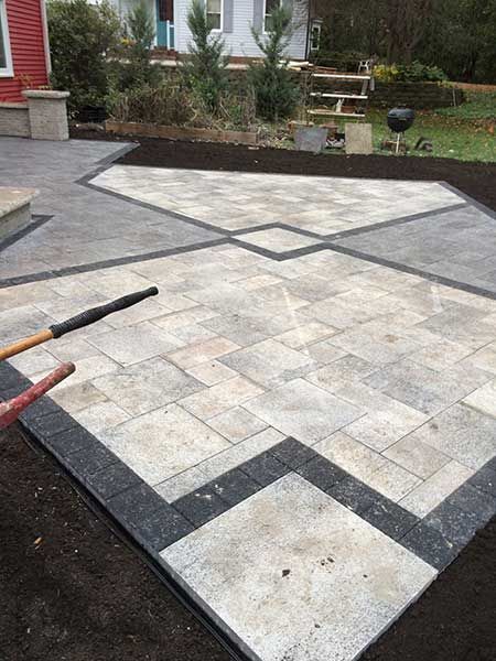 A patio paved with rectangular light gray stones framed by dark gray border bricks, set against a house and lawn.