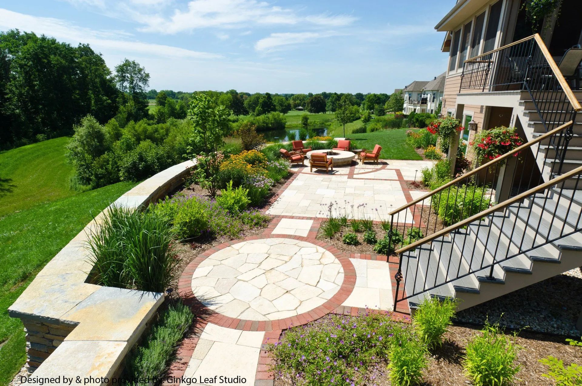 A patio with a stone fire pit and seating area, stone pathways, and landscaping, overlooking a grassy field and trees.