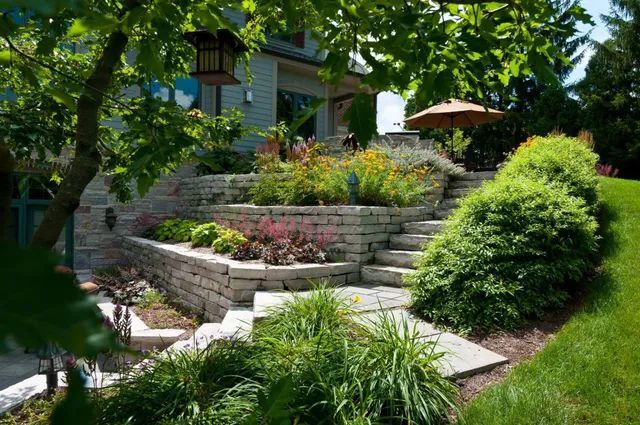 Tiered stone retaining walls and stairs landscaped with varied plants and shrubs lead up to a house with a patio umbrella.