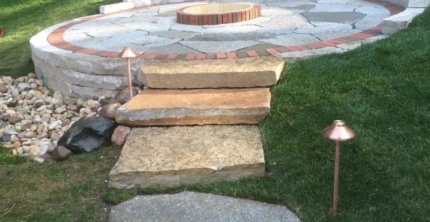 Three stone steps lead up to a raised circular patio with a central brick fire pit, flanked by two small garden lights.