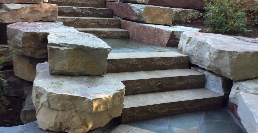 Natural stone staircase with rough-hewn boulders forming retaining walls and steps leading up through a garden.