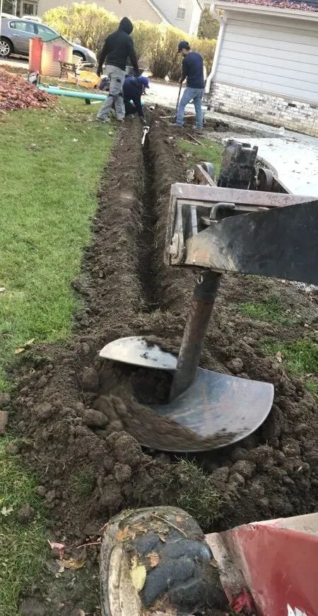 Workers use an auger to dig a narrow trench in a grassy residential yard near a house.