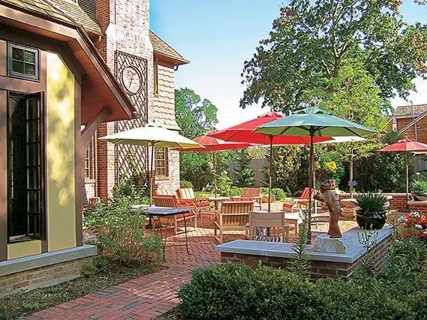 A brick patio with tables, chairs, and colorful umbrellas next to a house exterior on a sunny day.