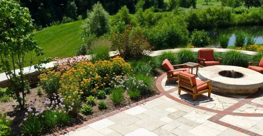 A patio with a round fire pit and four orange armchairs, overlooking a lush garden and a pond in a suburban backyard.