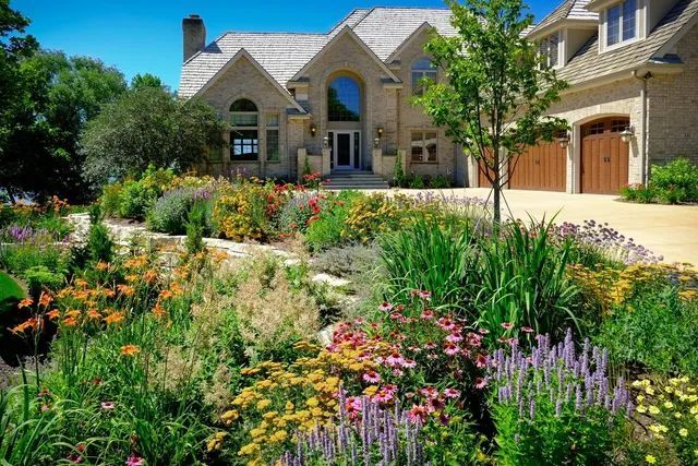 A large, multi-story stone house with a multi-car garage, fronted by a lush, vibrant garden of varied flowers and greenery.