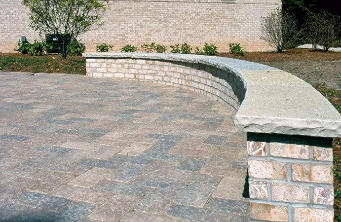 A curved stone seating wall made of tan bricks with a light grey stone cap, situated on a stone paver patio.
