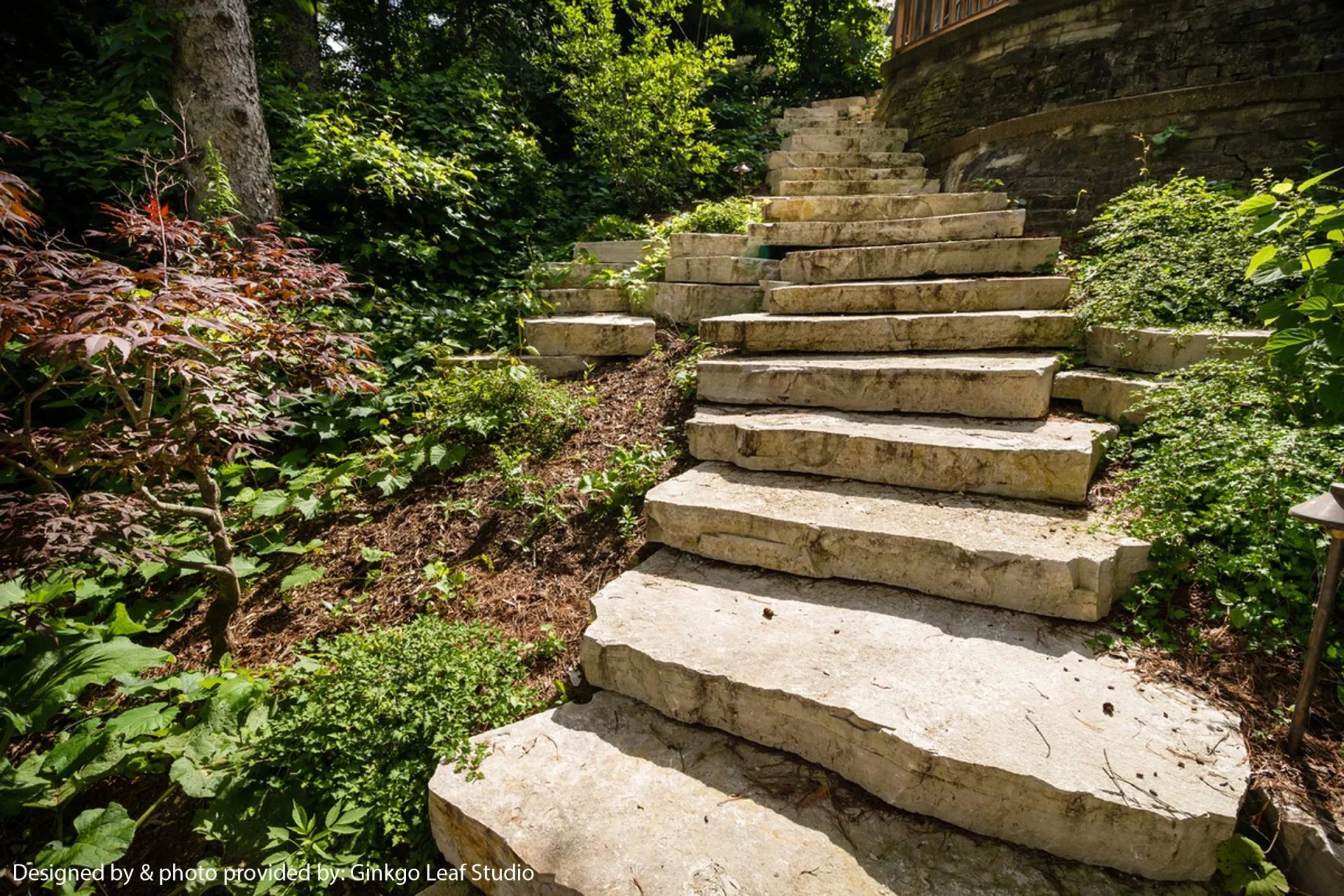 Stone steps set in a lush, green hillside landscape with dense foliage and a stone retaining wall.
