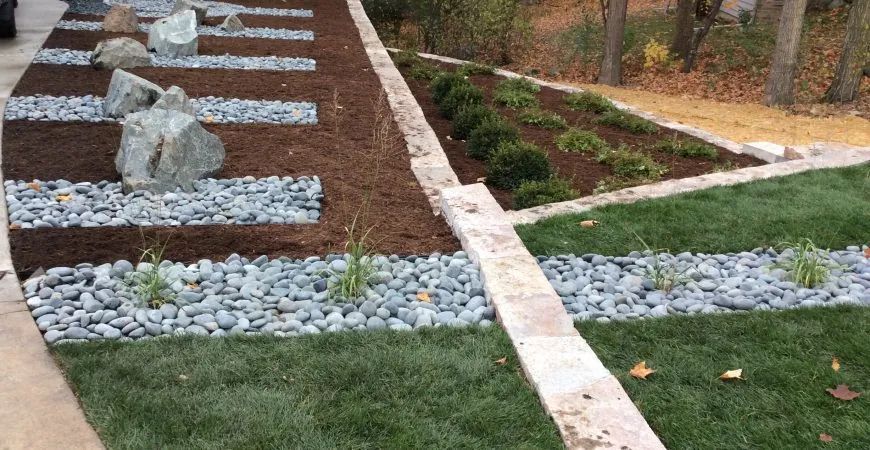 A modern landscape design featuring alternating stripes of river rocks, dark mulch, lawn, and stone retaining wall borders.