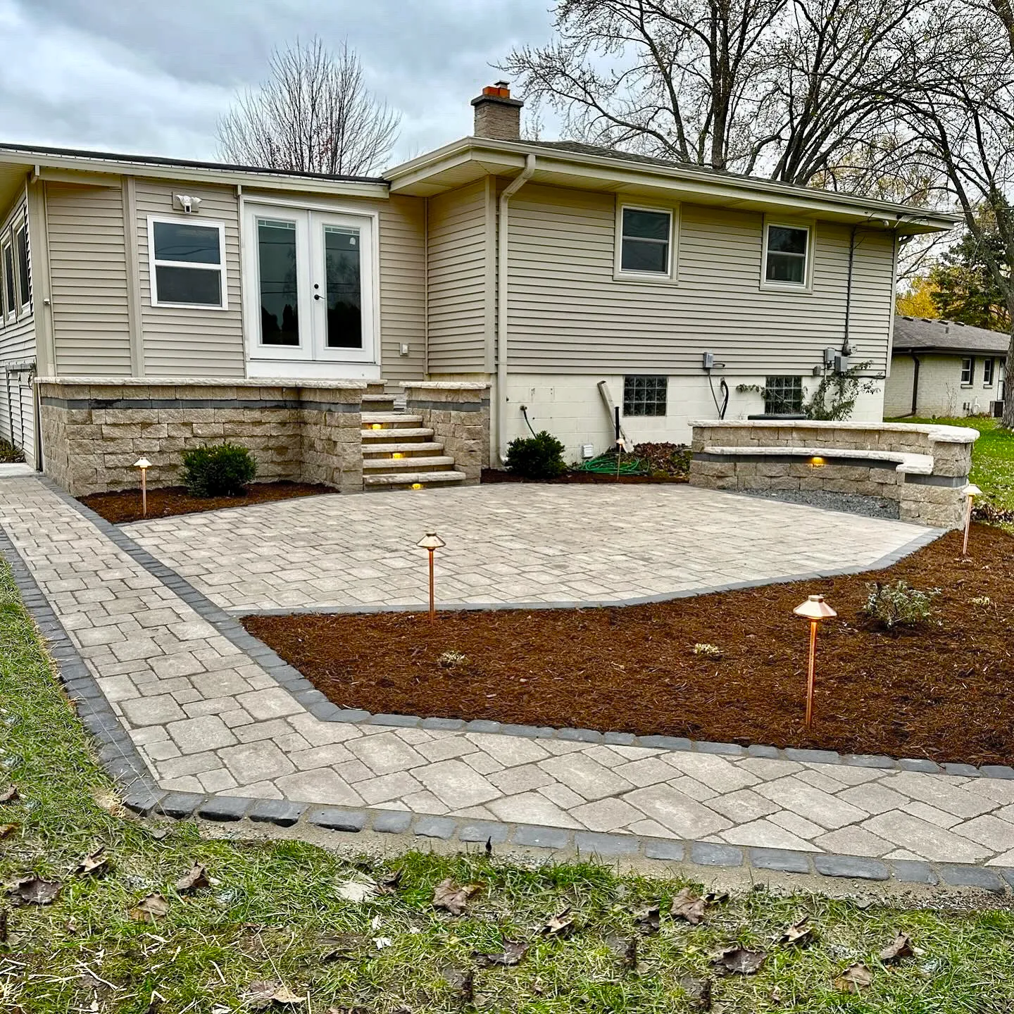 A beige house with a stone patio, a landscaped mulch bed, and a paved walkway with small garden lights in the yard.