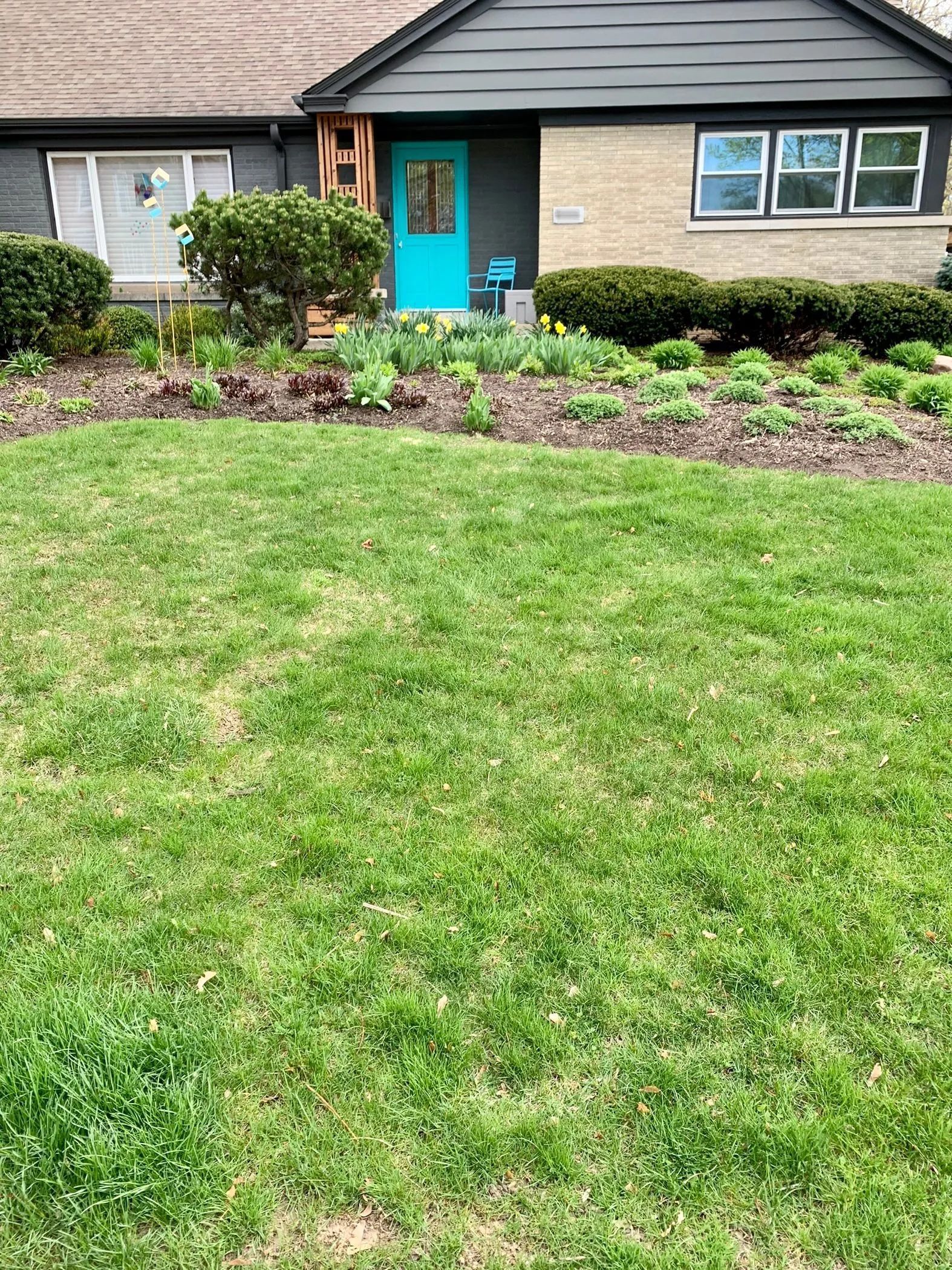 A house with light brick siding, a bright teal front door, dark roof, and a large front lawn with garden beds.