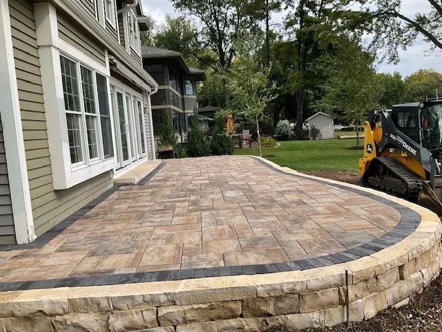 A new stone patio with a curved retaining wall extending from a house, with a small yellow construction vehicle nearby.