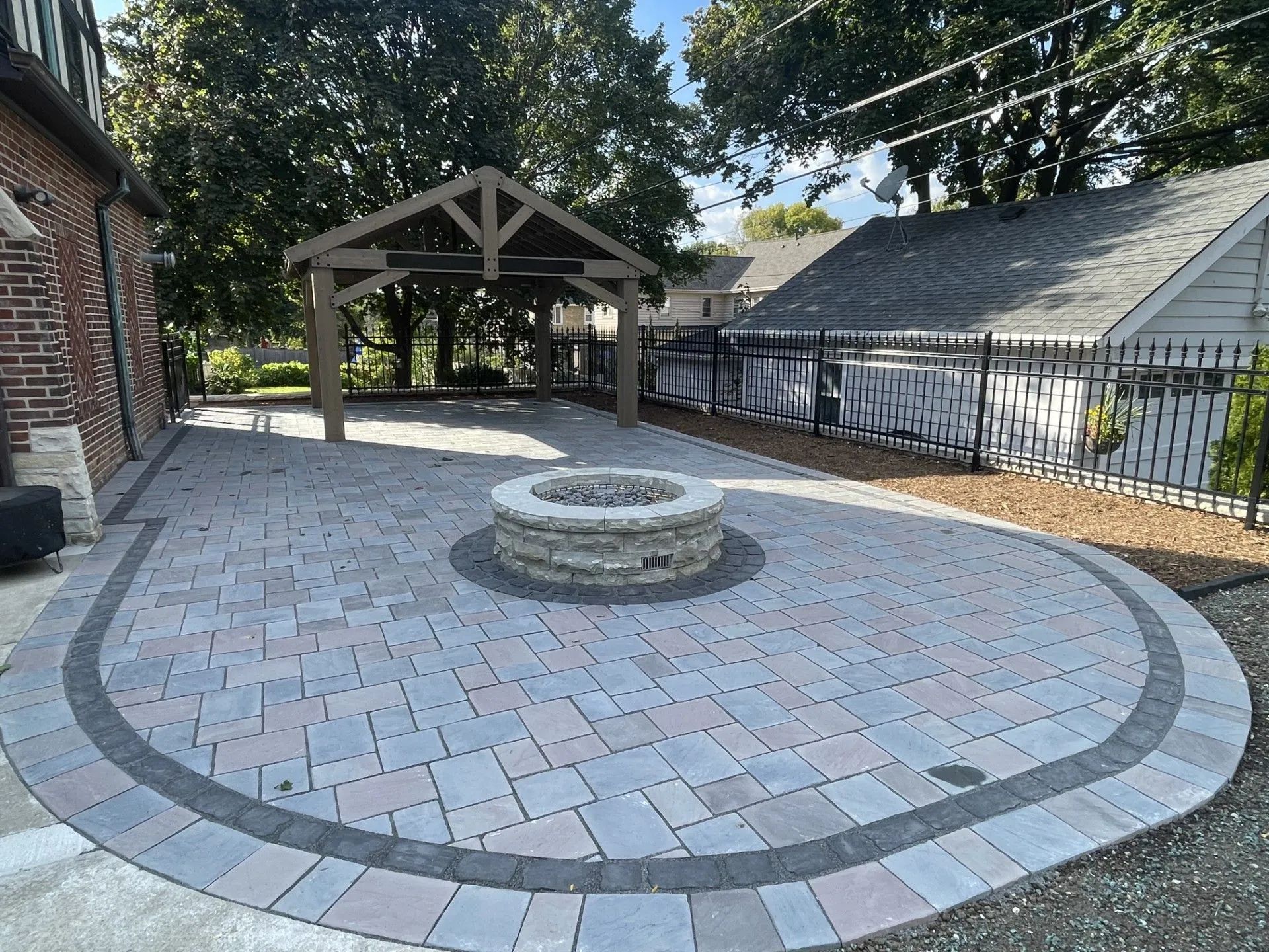A stone patio with a central circular fire pit and a wooden pavilion, set in a backyard next to a brick house.