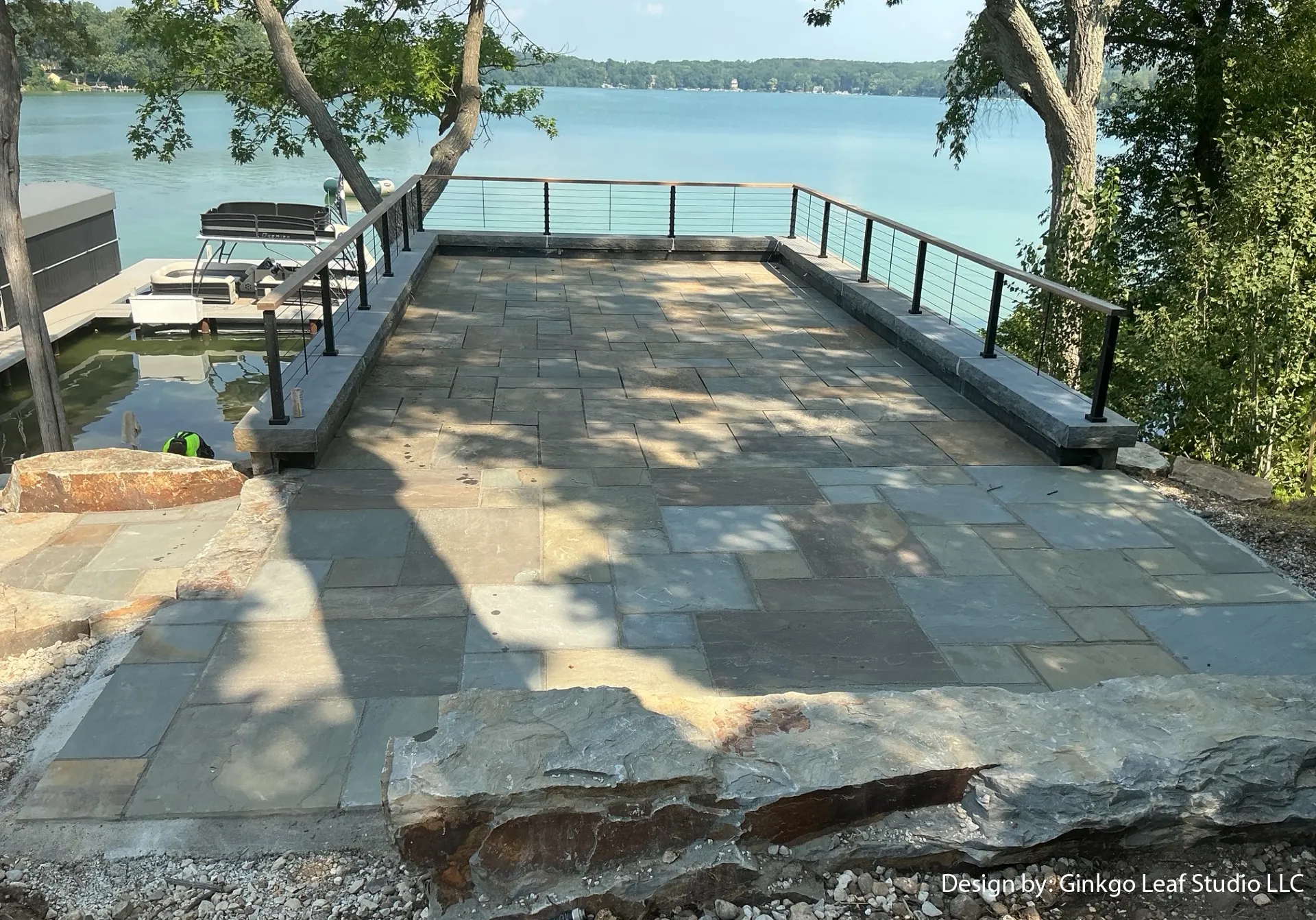 A rectangular stone patio overlooking a lake, featuring stone walls, metal railings, and a view of a wooden boat dock.