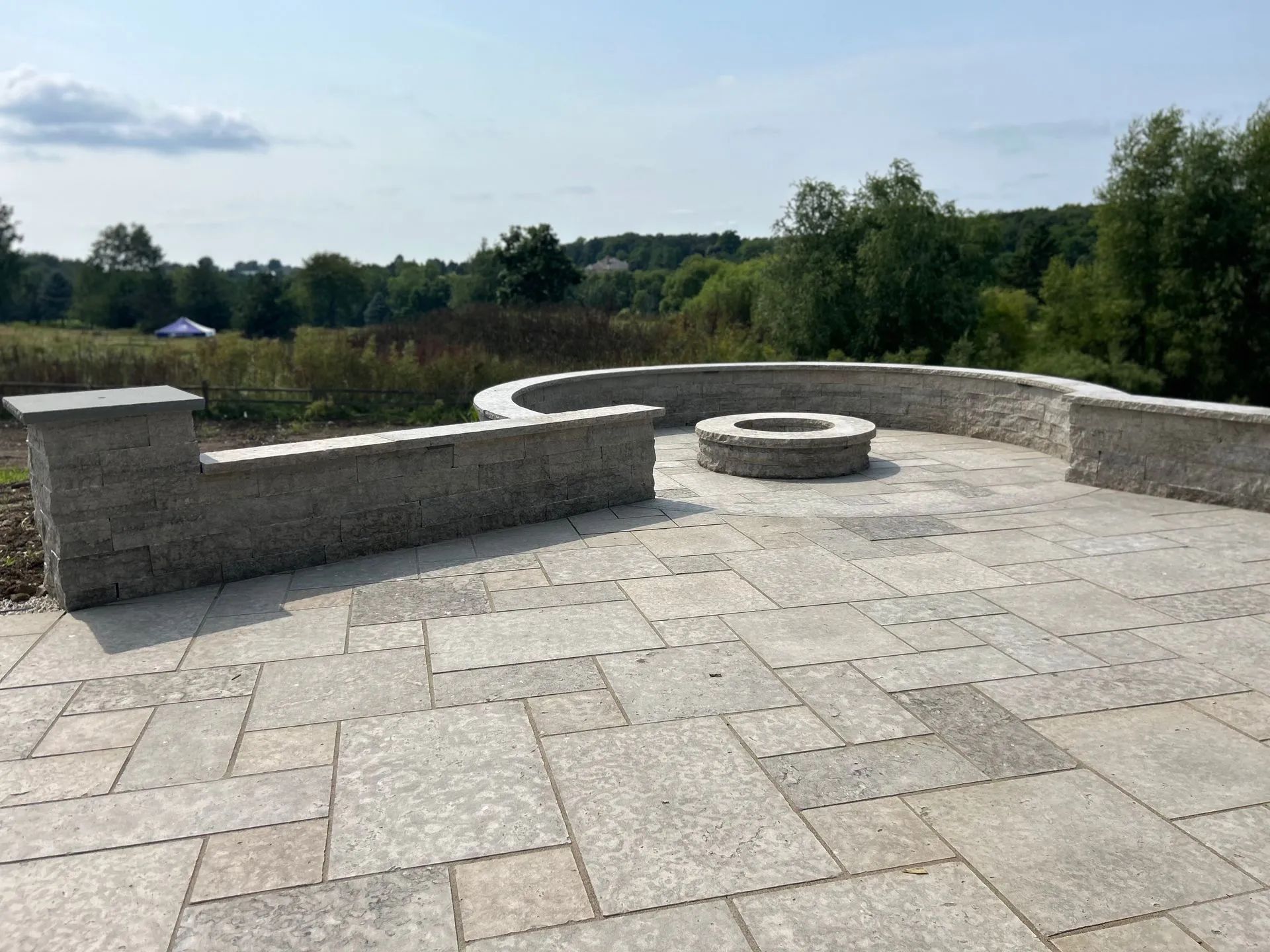A stone patio with a built-in circular fire pit, enclosed by a low stone retaining wall, overlooking a grassy landscape.
