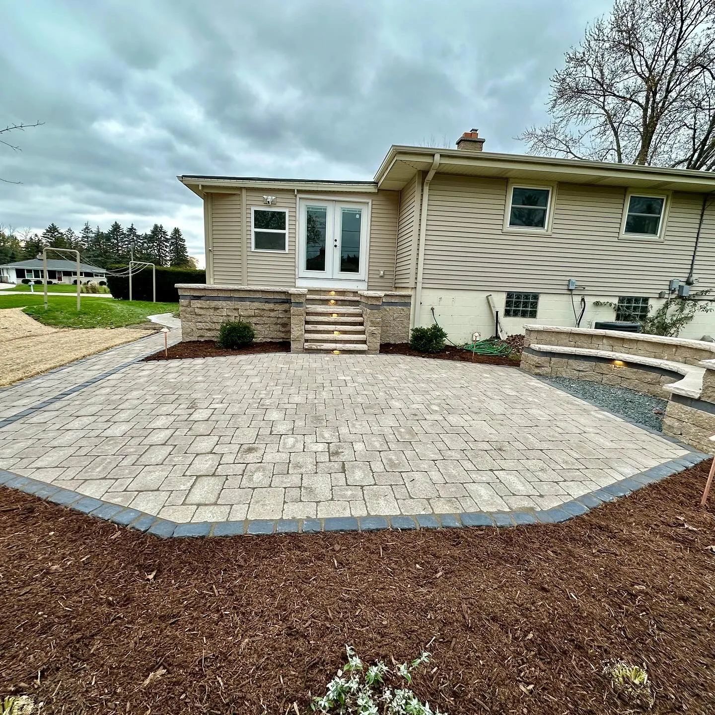 A new light-colored paver patio with a dark border sits in front of a house with beige siding and a stone staircase.