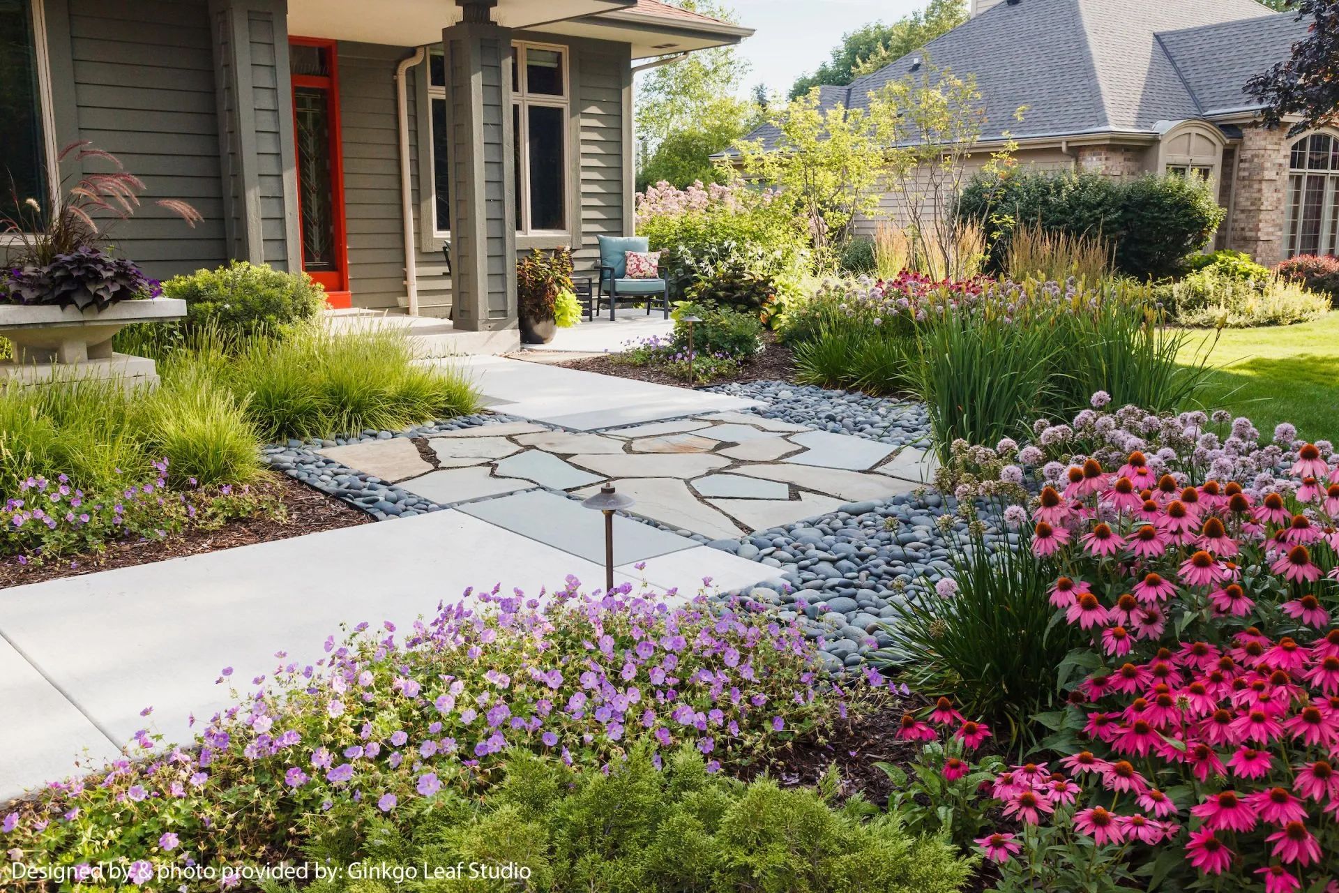 A stone pathway leads to a home entryway, framed by vibrant flower beds, greenery, and a decorative stone patio medallion.