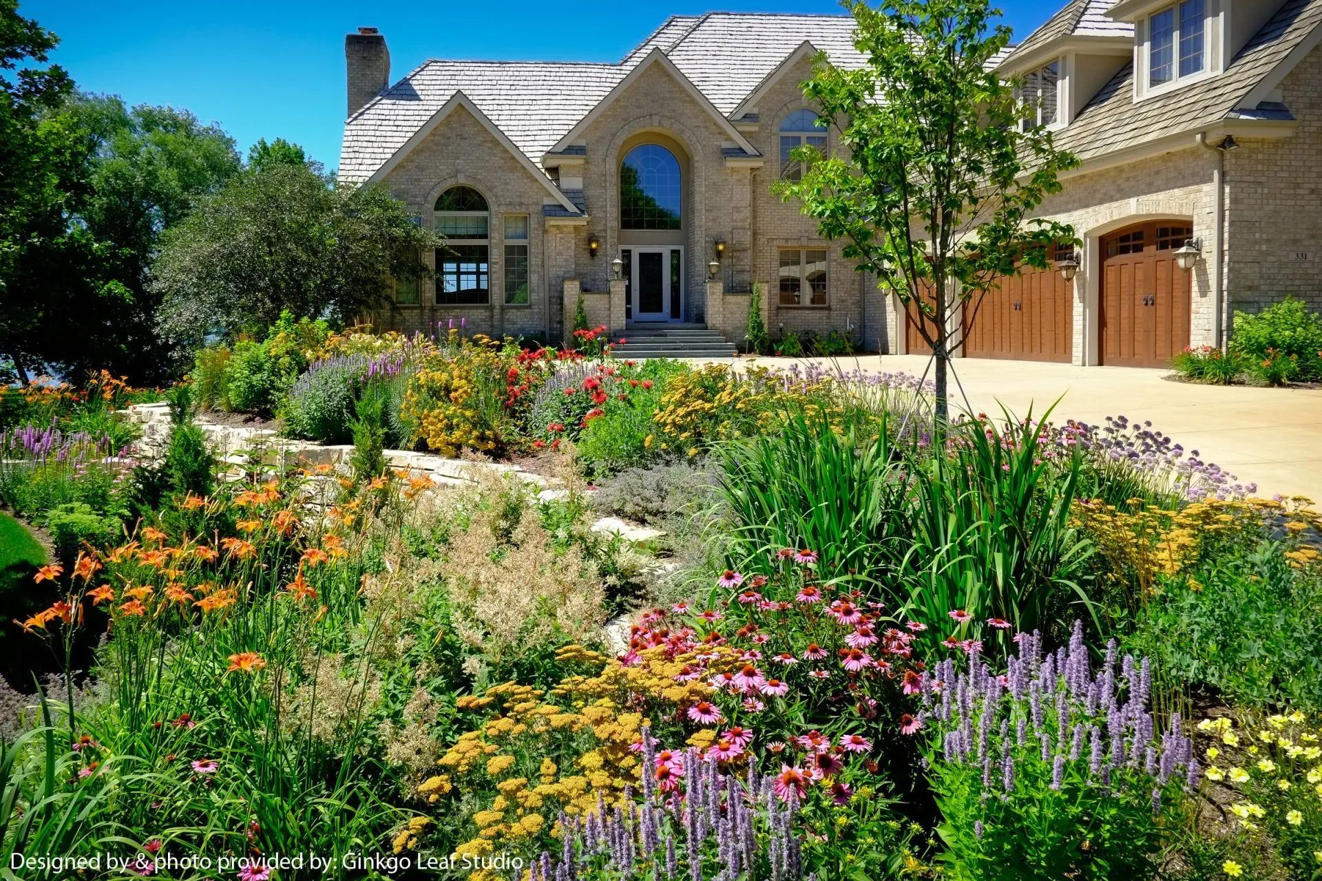 A large stone house with a multi-car garage overlooks a vibrant, multi-colored flower garden on a sunny day.