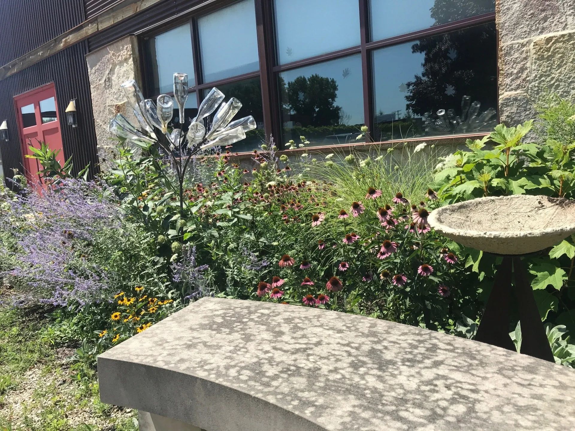 A stone bench sits before a garden bed with flowers, a birdbath, and a metal tree sculpture holding glass bottles.