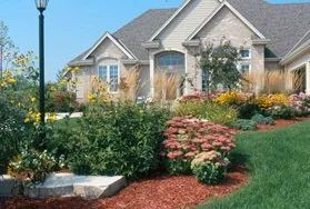 A suburban house with a tan brick exterior, a grey roof, and a front yard featuring shrubs, flowers, and a lamppost.