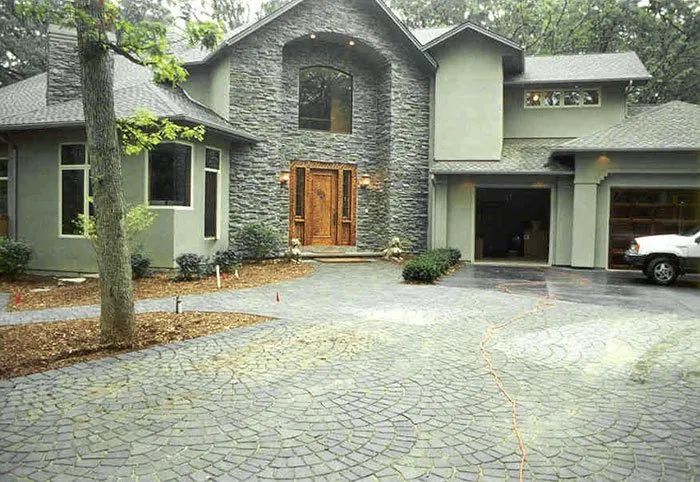 A two-story home with gray stone facade, stucco walls, and a patterned, paved circular driveway in a wooded setting.