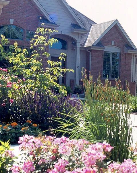 A brick house with a front porch, framed by a colorful garden with pink flowers, green foliage, and tall ornamental grass.