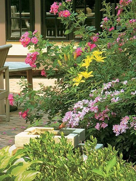A lush garden with pink and yellow flowers, a stone feature, and a patio with a chair near a house window.