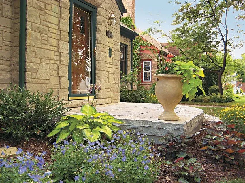 A stone front porch with a beige planter, surrounded by a lush garden, leads to the entrance of a rustic stone house.