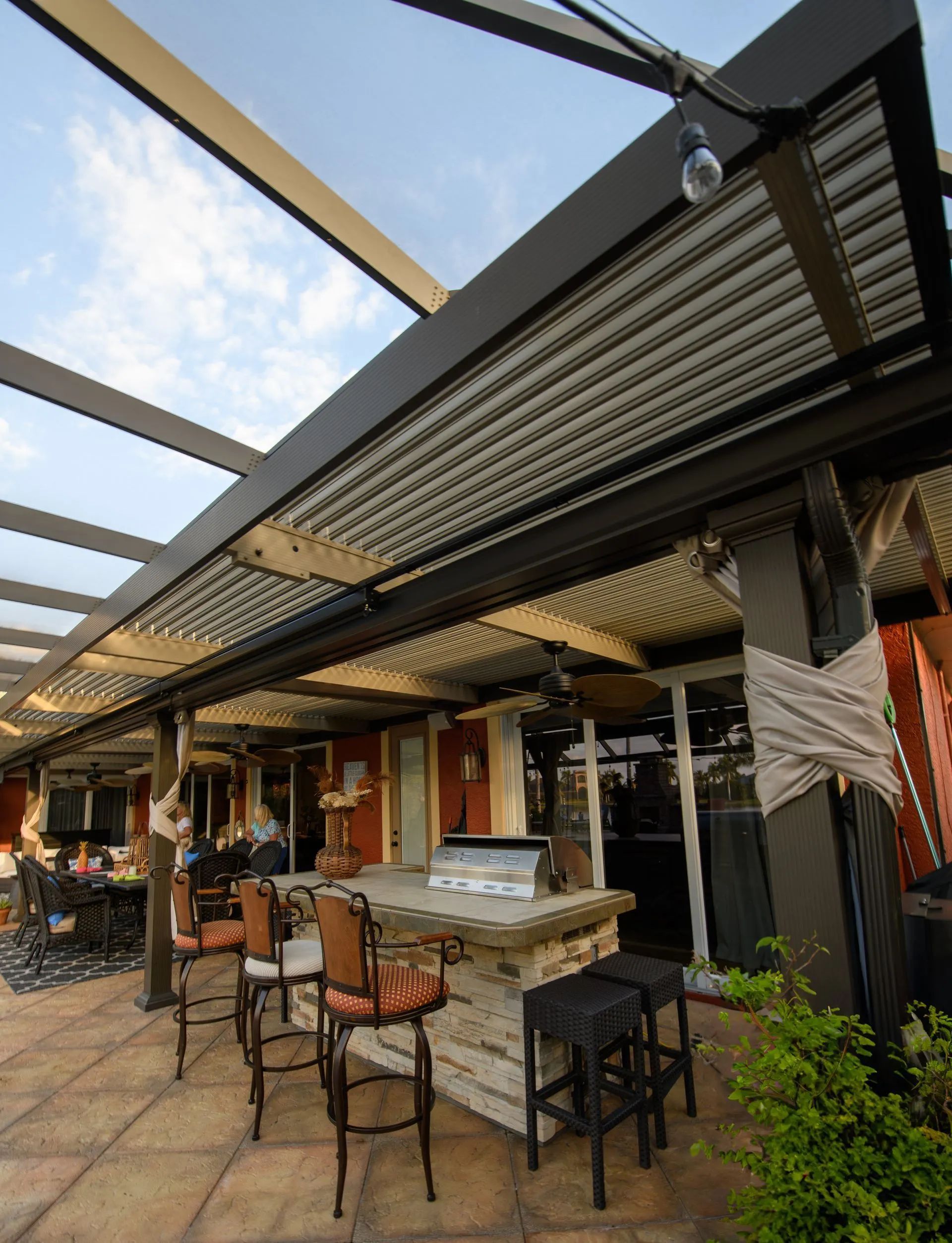 An outdoor patio featuring a stone bar with stools, a grill, and a corrugated metal roof, with dining area in the back.