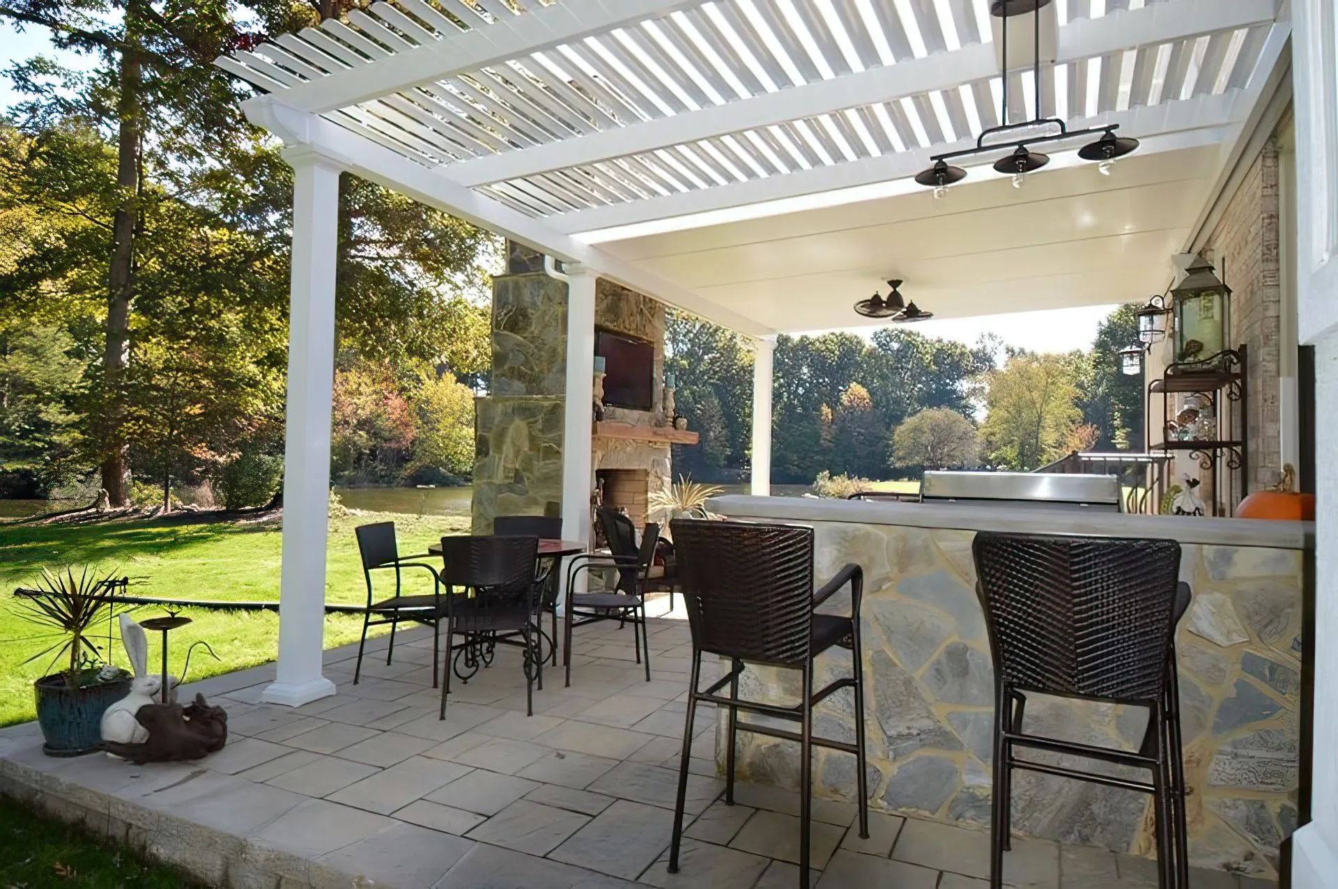 A stone patio under a pergola features an outdoor kitchen, bar seating, and a stone fireplace overlooking a grassy yard.