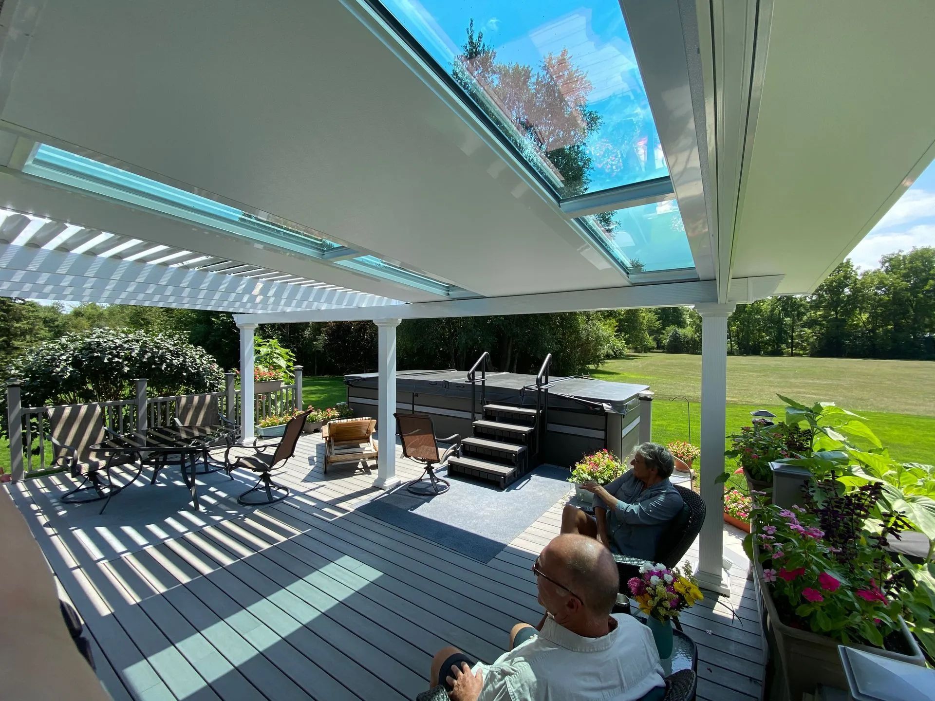 People relax on a grey deck under a pergola with retractable roof panels, overlooking a spa and grassy backyard.