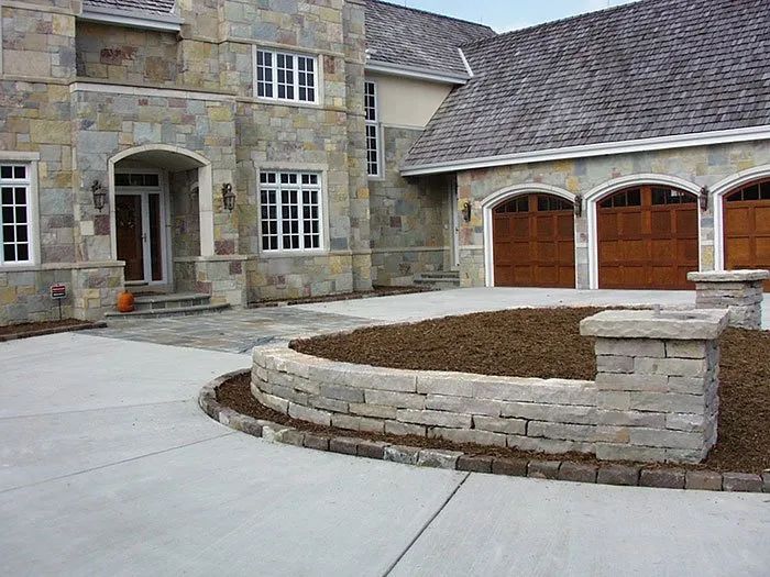 A stone house with a multi-car garage, featuring a curved stone retaining wall and a paved driveway in the foreground.