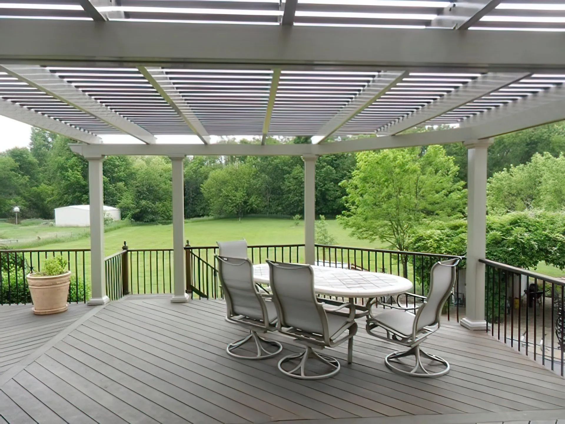 A furnished deck with a dining set beneath a pergola, overlooking a grassy yard with trees.