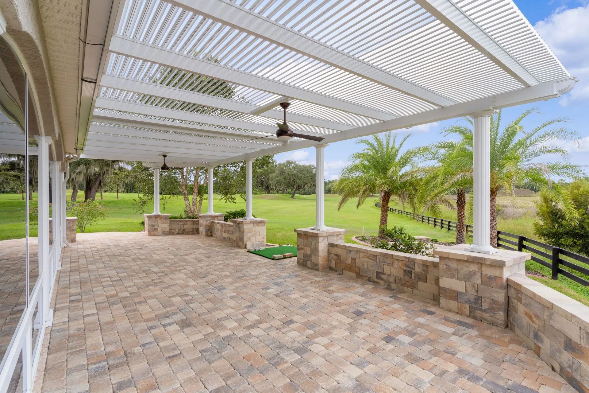 A covered outdoor stone patio with a ceiling fan, overlooking a grassy yard with palm trees and a black fence.
