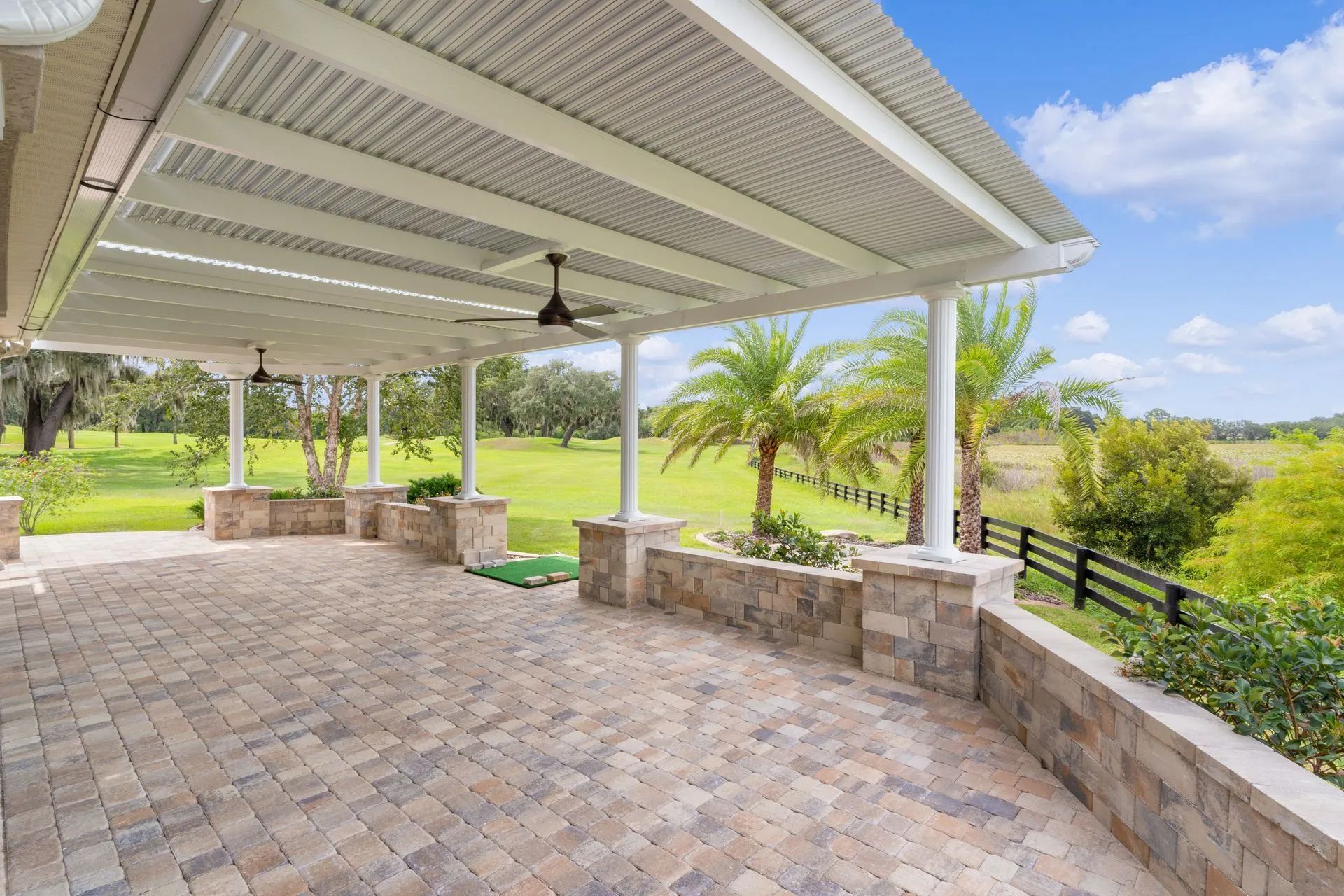 A paved patio with a metal roof, ceiling fans, and stone knee walls overlooking a grassy field and trees.