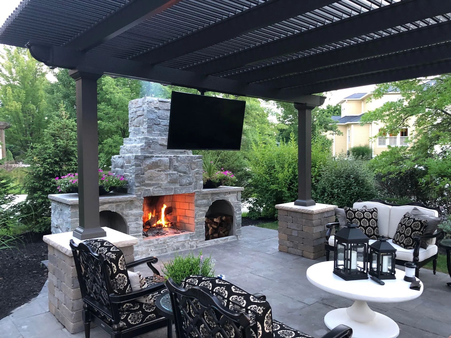 A stone outdoor fireplace with a hanging TV sits under a black pergola on a patio, with lounge chairs and a small table.