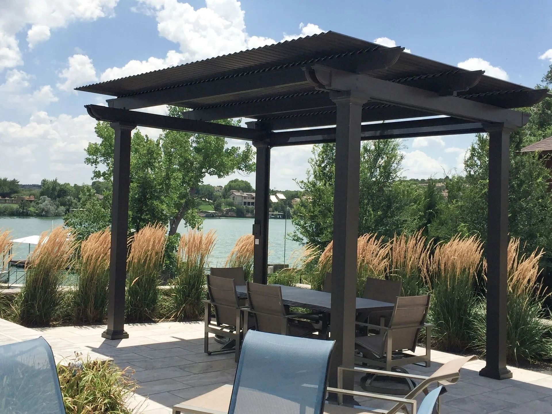 A dark metal pergola sits on a stone patio overlooking a lake, with a dining table, chairs, and tall grasses nearby.