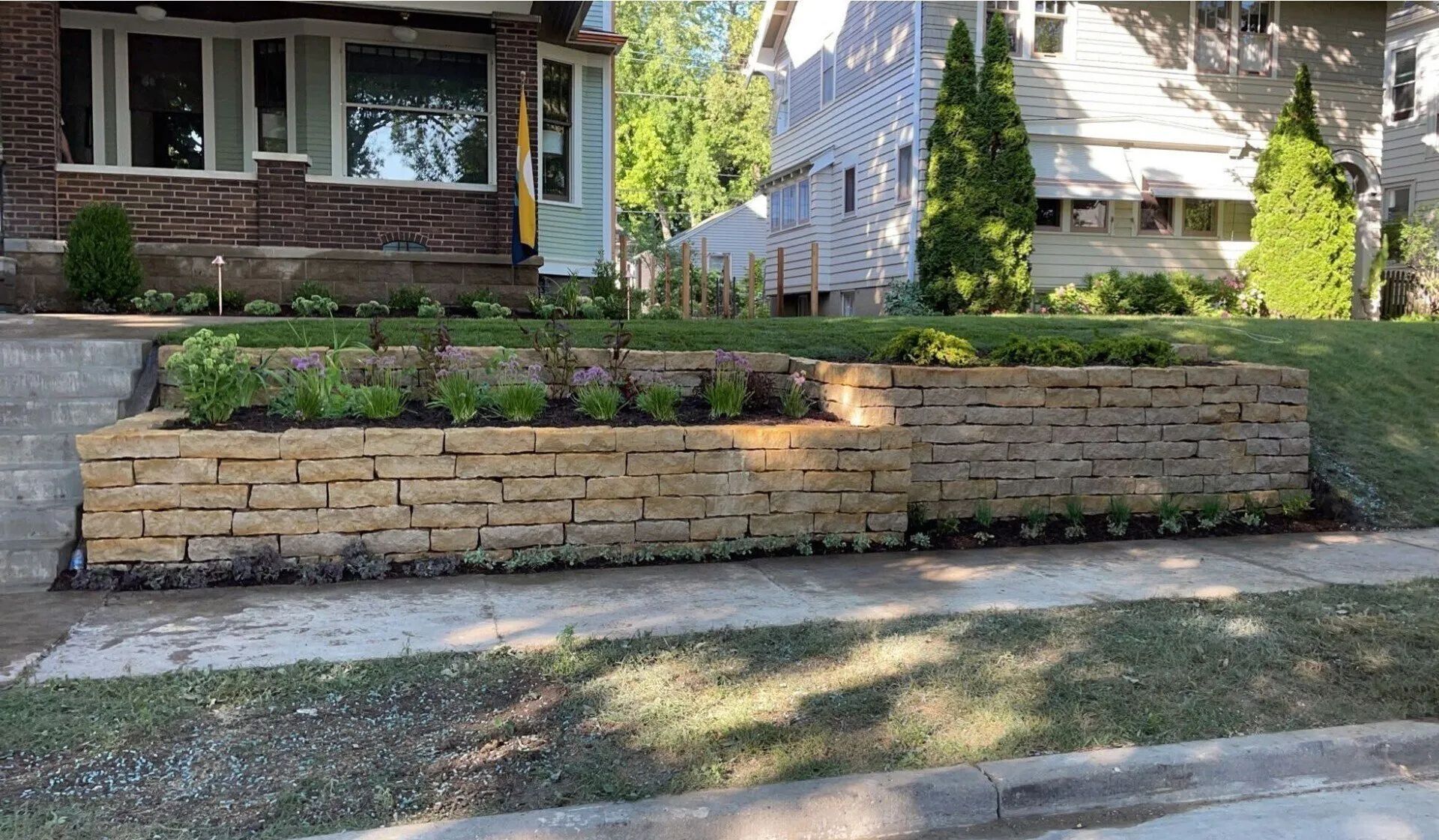 A tan stone retaining wall garden bed planted with greenery, situated in front of a residential home and sidewalk.