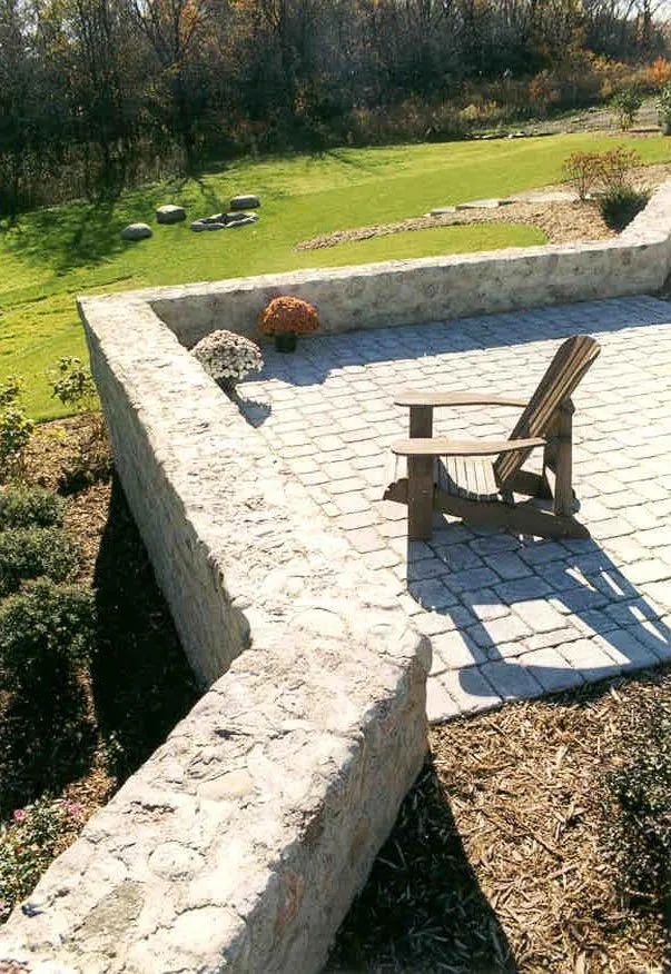 An Adirondack chair sits on a stone patio bordered by a low stone wall, overlooking a grassy yard with autumn foliage.