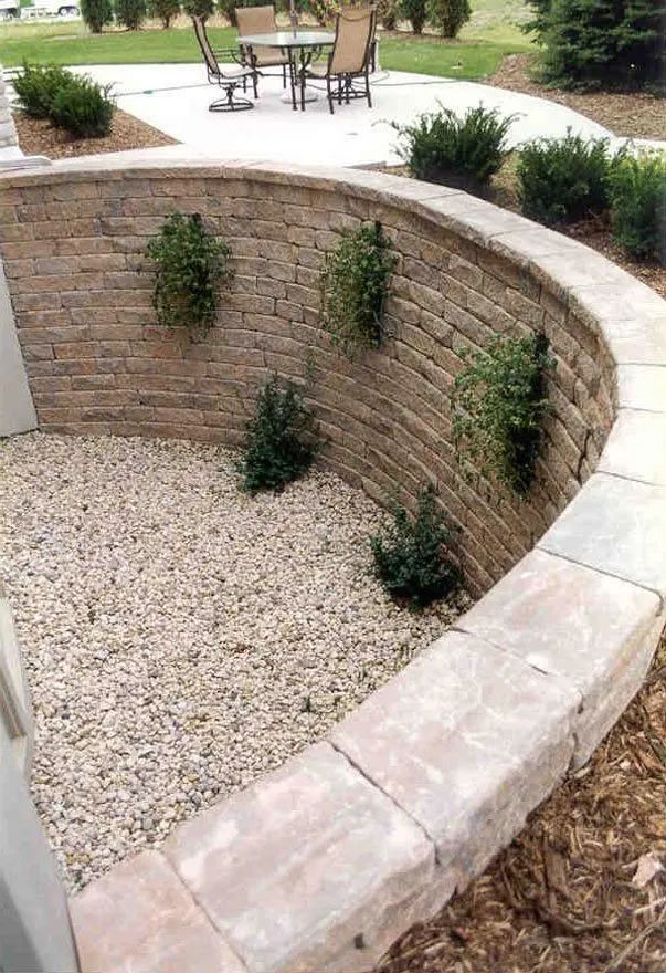 A curved, tan stone retaining wall creates a gravel-filled garden bed with small green shrubs on a patio next to a table.