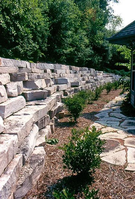 A tall, tiered natural stone retaining wall stands next to a flagstone path, mulch, and small green shrubs.