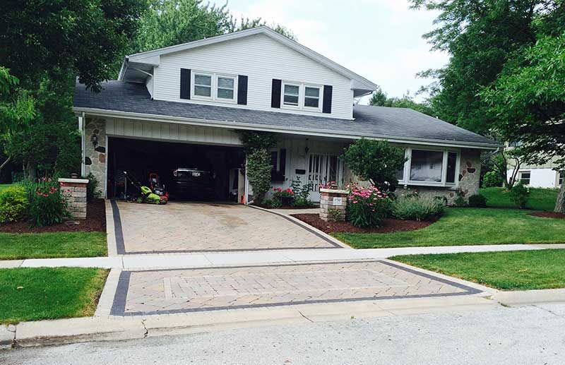 A two-story house with a white exterior, black shutters, and a decorative paver driveway surrounded by a green lawn.