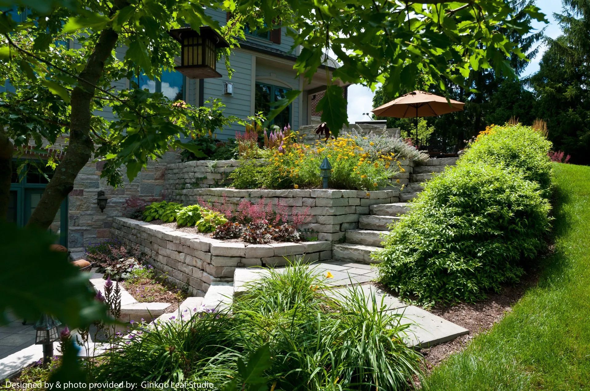 A tiered stone staircase leads to a patio beside a house, surrounded by lush green shrubs and colorful garden flowers.
