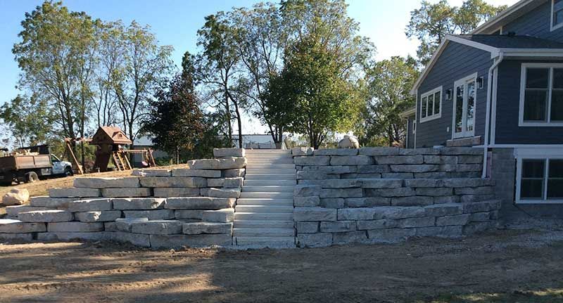 A backyard landscape featuring a stone retaining wall and central stone stairs leading up to a blue house.