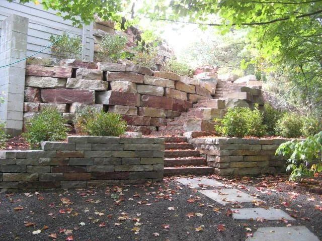 A tiered stone retaining wall with integrated steps leading up a hillside in a garden, scattered with autumn leaves.