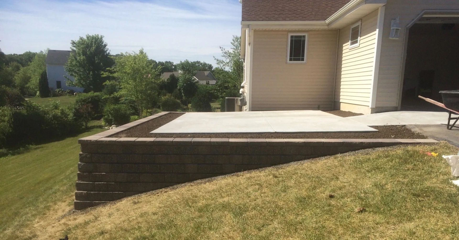A backyard features a raised concrete patio bordered by dark mulch and a stone retaining wall on a grassy slope.