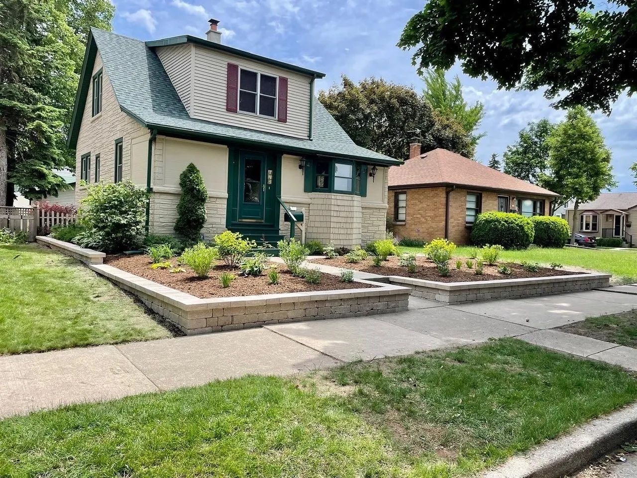 A light-colored house with a green roof features tiered stone retaining walls filled with young plants and mulch.