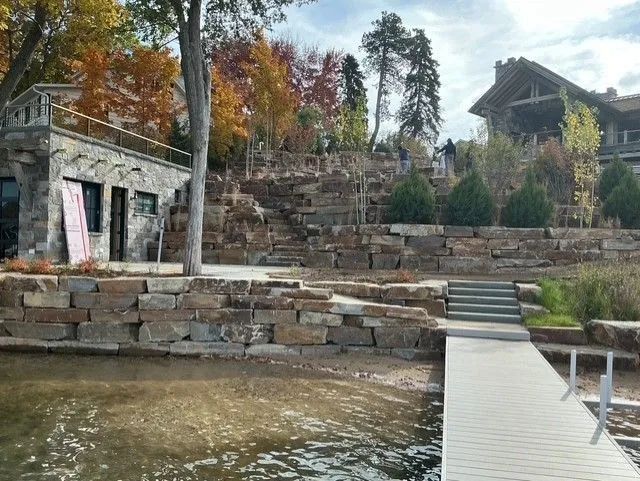 A stone building and tiered landscaping with a wooden dock extending into a lake under a bright, cloudy sky.